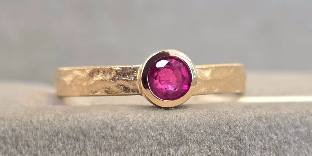 Goldring mit einem runden, pinken Rubin in der Mitte, auf einer grauen Oberfläche platziert.