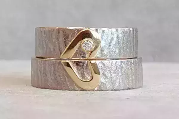 Zwei übereinanderliegende, selbst geschmiedete Trauringe mit strukturierter Oberfläche und einem sich ergänzenden Herzmotiv aus Gelbgold, verziert mit einem Brillanten.